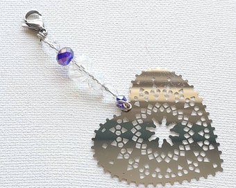 May include: A silver-colored heart-shaped charm with a decorative cut-out design. The charm is attached to a silver chain with clear and purple beads and a clasp. The charm is likely used as a pendant or a decorative accessory.