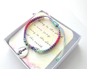 May include: A colorful macrame bracelet with a silver charm that says "PRAISE!" and a treble clef. The bracelet features pink, purple, and teal threads, and is presented in a white box with a card that has text that says "Sing to the Lord, a new song; sing to the Lord, all the earth."