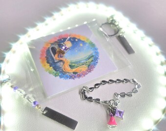 May include: A silver-toned chain bracelet with heart-shaped links, a purple square crystal charm, and a small pink bell charm. Included is a square card featuring a colorful illustration of a long-haired figure in a tower and a metal tag.