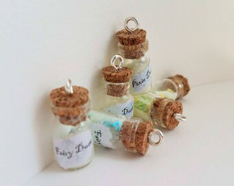 May include: Small glass bottles with cork stoppers, labeled "Fairy Dust" and "Pixie Dust." The bottles contain colorful glitter and small objects. Each bottle has a silver loop attached to the cork for hanging.