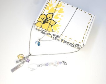 May include: A silver necklace with multiple charms, including a key, a purple gem, and a rectangular tag. A second necklace with blue and clear beads is also present. A white box with a yellow flower design and the text "Now's when my Life Begins" is in the background.