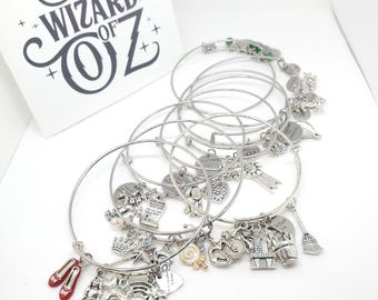 May include: Silver charm bracelets with various charms, including ruby slippers, a girl, a rainbow, and a castle. A white card with the text "The Wizard of Oz" is in the background. The bracelets are on a white surface.