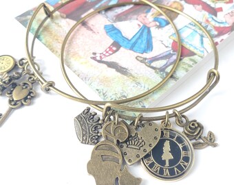 May include: Two antique brass-toned charm bracelets with Alice in Wonderland-themed charms. Charms include a clock, key, crown, heart, and a silhouette of Alice. A colorful illustration of Alice in Wonderland is in the background.