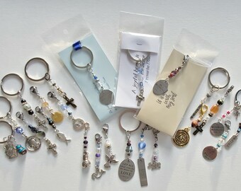 May include: A collection of keychains with various charms, including crosses, fish, and religious medallions. Some keychains have beaded accents in colors like blue, purple, and gold. Several keychains are packaged with cards that have text.
