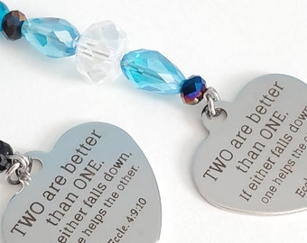 May include: Two heart-shaped pendants with the text "TWO are better than ONE" and a Bible verse. The pendants are silver-colored and connected by a strand of blue and clear beads. The beads have a faceted design.
