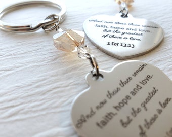 May include: A silver heart-shaped keychain with the engraved text "And now these three remain: faith, hope and love, but the greatest of these is love. 1 Cor 13:13." The keychain has a silver ring and a decorative amber-colored bead.