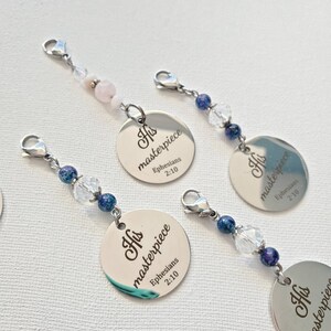 May include: Silver-toned charm with the inscription "His masterpiece Ephesians 2:10". The charm is attached to a chain with beads in shades of blue, clear, and pink. The charms have a lobster clasp.