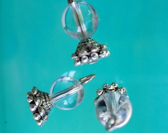 May include: Three clear glass and silver-toned metal bead charms. Each charm features a clear, spherical bead attached to a decorative silver-toned metal base and a small loop. The charms are displayed against a teal background.