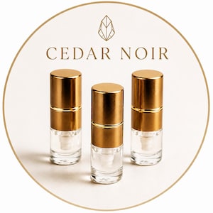May include: Three small, clear glass spray bottles with metallic gold caps arranged in a cluster. Above the bottles, the text CEDAR NOIR is printed in gold, accompanied by a geometric, stylized leaf or crystal icon centred above the text.