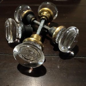 May include: Two sets of clear glass door knobs with brass-toned metal bases and central metal spindles. The knobs feature a smooth, rounded profile and are arranged in a crossed configuration, showcasing the reflective glass and metallic hardware components.