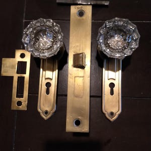 May include: A set of two clear, faceted glass door knobs attached to brass backplates with keyholes, accompanied by a central brass mortise lock mechanism and a separate brass strike plate, all featuring a metallic finish and clear glass texture.