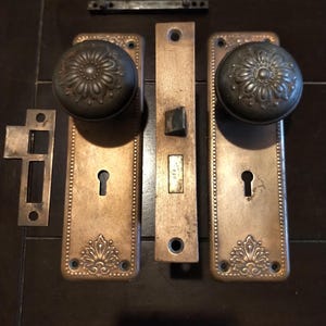 May include: A set of antique brass door hardware including two rectangular backplates with floral embossed details, two round matching knobs, a central latch mechanism, a strike plate, and a small metal bar, all featuring a weathered patina.