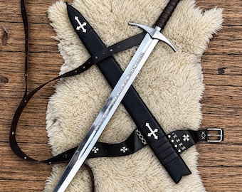 May include: A silver-toned steel sword with a dark brown leather-wrapped grip and a circular pommel featuring a red cross emblem. The blade is etched with text and rests alongside a black leather scabbard adorned with silver cross accents.