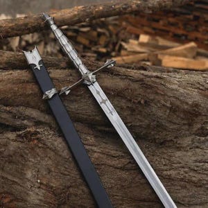 May include: A silver-toned metal sword featuring a ribbed handle, a cross-shaped guard, and a blade with an etched decorative pattern. The sword is accompanied by a long, black scabbard with silver-colored metal fittings at the top and middle.