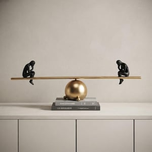 May include: A decorative sculpture featuring two matte black human figures in a seated, contemplative pose, positioned on opposite ends of a long, horizontal gold-toned rod balanced atop a central gold sphere resting on two stacked books.