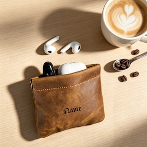 May include: A brown leather pouch with a top opening, featuring the word Name embossed in black gothic lettering. The pouch contains a white wireless earbud charging case and a black object, with two white wireless earbuds placed nearby.