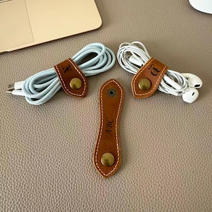 May include: Three tan leather cable organizers with white stitching and brass-colored metal snap buttons. Two organizers are wrapped around coiled white and light blue cables, while one is flat and features the embossed letters ABC in the center.