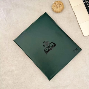 May include: A rectangular dark green folder with stitched edges. The front cover features a black engraved graphic of an open book topped with a compass rose, and the initials DM are engraved in the bottom right corner in a serif font.