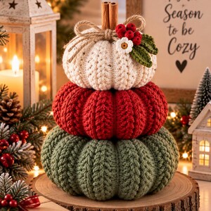 May include: A stacked pumpkin decoration featuring three crocheted pumpkins in cream, red, and green. The top pumpkin is adorned with a cinnamon stick stem, a jute twine bow, a small white flower, and red berries with green leaves.