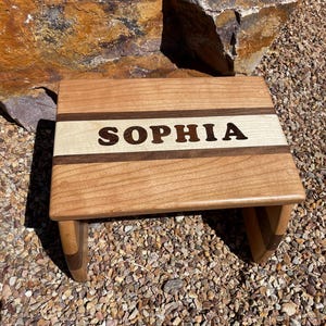 May include: A wooden step stool with a light brown finish. The top of the stool has a white stripe and the name "SOPHIA" is engraved in dark brown letters.