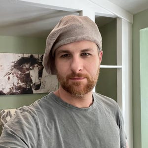 Op de afbeelding: Een lichtbruine gebreide baret. De baret is gemaakt van een textuurrijk materiaal en wordt op het hoofd gedragen. De persoon op de foto draagt een grijs t-shirt.