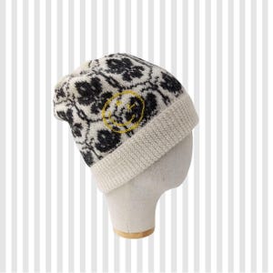 Op de afbeelding: Een gebreide beanie met een crèmekleurige rand en een zwart-crèmekleurig bloemenpatroon. Een gele geborduurde smiley staat op de zijkant. De hoed wordt getoond op een mannequinhoofd.