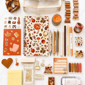 May include: A collection of stationery items including a spiral-bound notebook with a pumpkin and leaf pattern, a mesh pencil case, a brown scrunchie, various pens, stickers, sticky notes, gold paper clips, a candle, and a small white portable device.