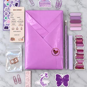 Op de afbeelding: Een paars ingepakt boek met een hartvormig zegel, vergezeld van briefpapier zoals een boekrecensiekaart, een potlood, een markeerstift, paarse paperclips, boekenstickers, een vlindersticker, een maansticker en een taartsticker.