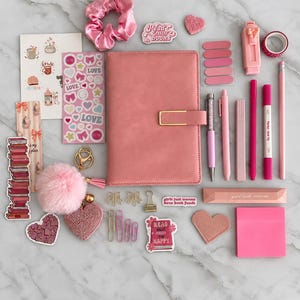 Puede incluir: Un conjunto de papelería rosa que incluye un cuaderno central con cierre dorado, rodeado por un coletero de satén, varias pegatinas con texto como Read more books, bolígrafos, rotuladores, un llavero de pompón, corazones y clips de papel.