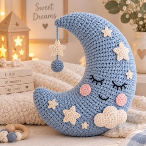 Può includere: Un cuscino all'uncinetto a forma di mezzaluna color azzurro, caratterizzato da un volto sorridente con occhi chiusi e guance rosa. Il cuscino è decorato con stelle e una nuvola all'uncinetto color crema, con una stella e una pallina blu pendenti.