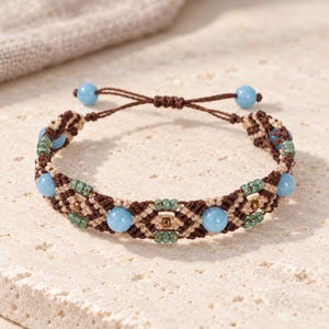 May include: A macrame bracelet featuring dark brown and beige knotted cord, accented with light blue round beads and small green seed beads. The adjustable closure includes two light blue beads at the ends of the brown cord ties.