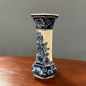 May include: A hexagonal ceramic vase featuring a white base with intricate blue floral patterns. The design includes a flared rim, a slender neck, and a wider base, all decorated with detailed blue blossoms and scrolling foliage motifs.