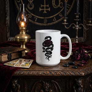 May include: A white ceramic mug featuring a black graphic illustration of a coiled snake wrapped around a dark red rose. The mug has a standard handle and a smooth finish, centered on a dark surface with other decorative items visible nearby.