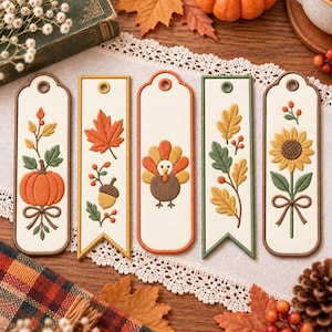 May include: Five rectangular bookmarks with embroidered autumn designs on a cream background. The designs include a pumpkin, an acorn with leaves, a turkey, oak leaves with berries, and a sunflower, each featuring a hole at the top.