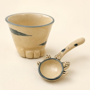 Puede incluir: Una taza de cerámica beige con borde azul oscuro y detalles en forma de hoja, con una base moldeada como una pata. Junto a ella hay una cuchara de cerámica beige a juego con mango largo, decorada con puntos azul oscuro y pequeñas orejas.
