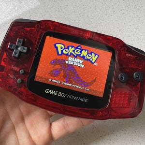 May include: A translucent red Game Boy Advance handheld gaming console featuring a black directional pad, start and select buttons, and A and B buttons. The screen displays the title screen for the video game Pokémon Ruby Version with text.