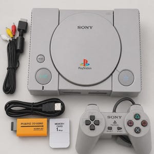 以下が含まれることがあります： グレーのソニーPlayStationゲーム機本体、同色のコントローラー、黒いAVケーブル、黒いHDMIケーブル、白い1MBメモリーカード、オレンジ色のPS1&PS2 HDMI変換アダプター。本体にはディスクカバー、電源、リセット、オープンボタンが備わっています。