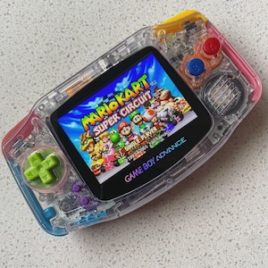 Può includere: Una console portatile Game Boy Advance trasparente con il gioco Mario Kart Super Circuit sullo schermo. Il dispositivo presenta una croce direzionale verde brillante, pulsanti azione rossi e blu e accenti colorati lungo i bordi.