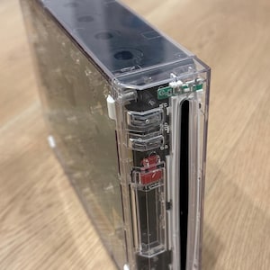 Könnte beinhalten: Eine rechteckige Videospielkonsole mit einem transparenten, durchsichtigen Kunststoffgehäuse, das die internen elektronischen Komponenten sichtbar macht. Die Vorderseite enthält einen vertikalen Disc-Schacht, einen roten Netzschalter und einen schwarzen Innenrahmen.