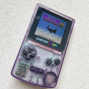 以下が含まれることがあります： 半透明の紫色のゲームボーイカラー携帯ゲーム機。画面には「ポケットモンスター 金」のタイトル画面が表示されており、空飛ぶ生き物のシルエット、黒い十字キー、Aボタン、Bボタン、セレクトボタン、スタートボタンが配置されています。