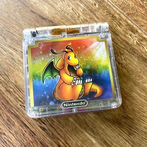 以下が含まれることがあります： 透明なプラスチック製の携帯ゲーム機で、カバーにはオレンジ色のカイリューのキャラクターが鮮やかに描かれています。本体は正方形の折りたたみ式で、黒い楕円形の背景に白いNintendoのロゴがプリントされています。