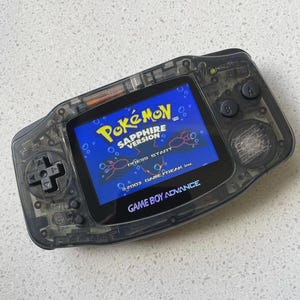 Può includere: Una console portatile Game Boy Advance nera trasparente con uno schermo retroilluminato che mostra la schermata del titolo di Pokémon Sapphire Version, con il testo Press Start e 2003 Game Freak inc. sul display blu brillante.