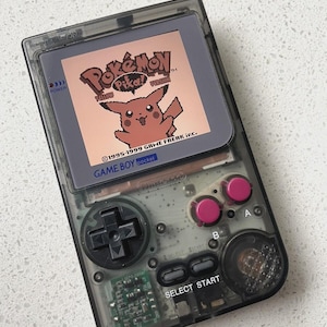 以下が含まれることがあります： 透明な黒色のゲームボーイポケット携帯ゲーム機。画面にはピカチュウが描かれた「ポケットモンスター ピカチュウ」のタイトル画面が表示されている。本体には黒い十字キー、2つのマゼンタ色のボタン、黒いセレクトボタンとスタートボタンが配置されている。
