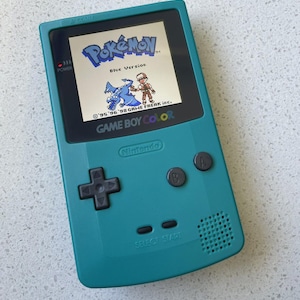 May include: A teal Game Boy Color handheld gaming console featuring a screen displaying the Pokémon Blue Version title screen with a character and dragon graphic, a black directional pad, A and B buttons, and Select and Start buttons.