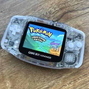 Op de afbeelding: Een doorzichtige Game Boy Advance handheld spelcomputer met een wit D-pad en witte knoppen. Het scherm toont de titel Pokémon Liquid Crystal Version met een afbeelding van een blauw wezen in een landschap met kliffen.