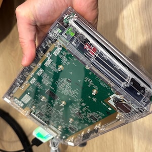 Könnte beinhalten: Ein elektronisches Gerät aus transparentem Kunststoff mit einer grünen Leiterplatte und sichtbaren internen Komponenten, darunter eine kleine runde Batterie, Metallanschlüsse und ein Schlitz, die komplexe Schaltkreise zeigen.