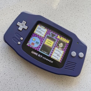 Può includere: Una console portatile Game Boy Advance di colore viola con un pad direzionale grigio chiaro, pulsanti A e B, e pulsanti Start e Select. Lo schermo mostra l'interfaccia del gioco Dr. Mario con un messaggio di Game Over, punteggio e livelli.