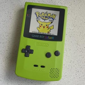 May include: A lime green Game Boy Color handheld gaming console featuring a screen displaying the Pokémon Yellow Version title screen with Pikachu. The device includes a black directional pad, A and B buttons, Select and Start buttons, and a speaker.