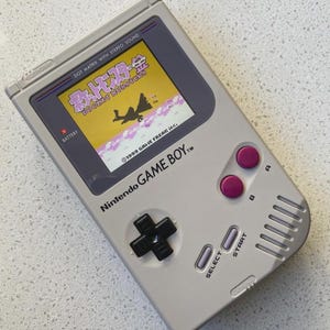 以下が含まれることがあります： 黒い十字キー、2つの紫色のAボタンとBボタン、灰色のセレクトボタンとスタートボタンを備えたライトグレーの任天堂ゲームボーイ携帯ゲーム機。画面には日本語のポケットモンスター金ゲームタイトルとロゴが表示されています。