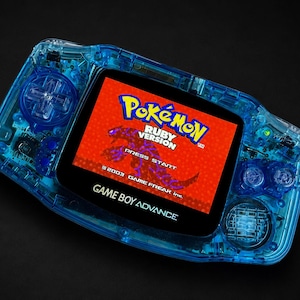 Può includere: Una console portatile Game Boy Advance blu traslucido con uno schermo luminoso che mostra la schermata del titolo di Pokémon Ruby Version, con il testo Pokémon Ruby Version, Press Start e 2003 Game Freak inc visibile sul display.